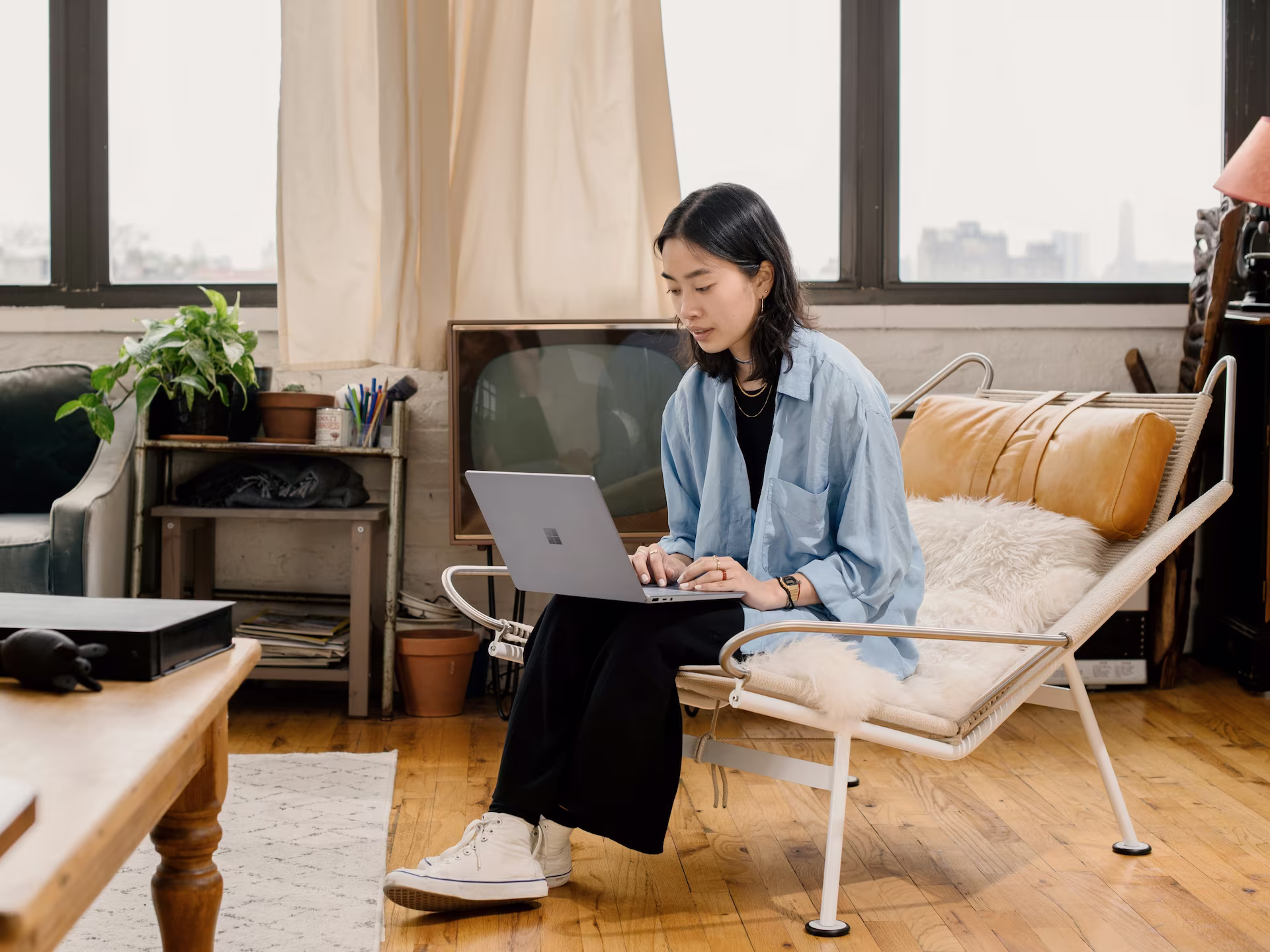 A young woman leaning over a table, writing in a notebook with an open laptop in front of her in a cozy indoor setting.