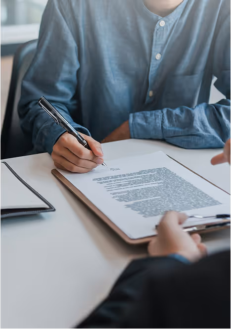 A close-up photograph showing a person in a blue shirt signing a document with a pen.